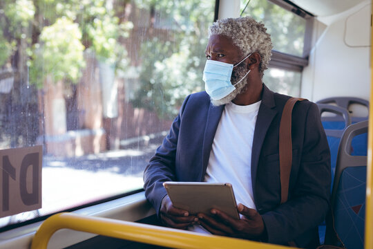 African american senior man wearing face mask sitting on bus using digital tablet looking out of win