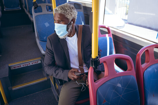 African american senior man wearing face mask and earphones sitting on bus holding smartphone