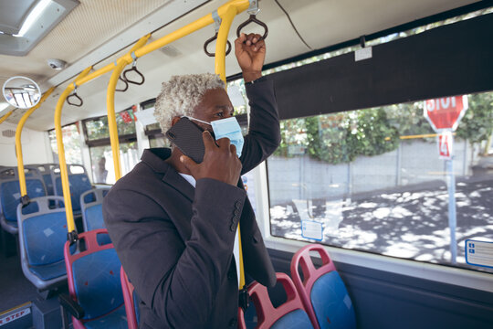 African american senior man wearing face mask standing on bus talking on smartphone