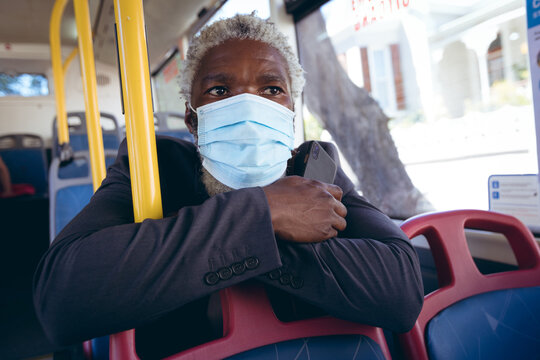 African american senior man wearing face mask sitting on bus holding smartphone - Powered by Adobe