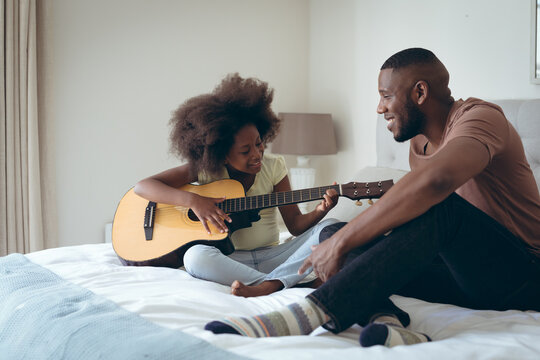 African american man and his daughter sitting on bed girl is playing guitar