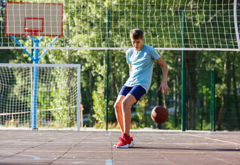 Cute Teenager in green t-shirt with orange basketball ball plays basketball on street playground in summer. Hobby, active lifestyle, sports activity for kids.	