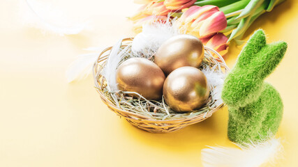 Golden easter colour eggs in basket with spring tulips, white feathers on pastel yellow background in Happy Easter decoration. Spring holiday concept.