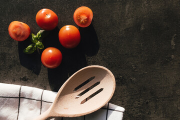 Red cherry tomatoes, green basil, wooden spoon, towel on gray background