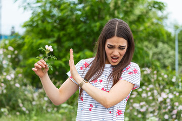 Young woman with Pollen allergy holding a flower and saying no.. Young woman with pollen and grass allergies. Flowering trees in background. Spring Seasonal allergies and health problems.