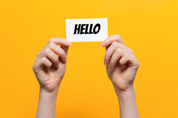 Woman's hands hold a white card with the inscription Hello. Close up. Yellow background. The concept of the world hello day