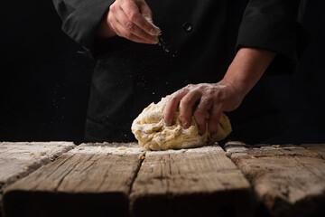 The dough in the hand of the chef on a dark black wood background. Baking bread, pizza, pasta. A recipe from the chef for pizza, bread or pastries. Home kitchen with space for text for menu design