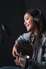 Obraz na płótnie Canvas Asian female singer with a passion for music and microphone. While playing her guitar in a professional studio. Music concept, sound recording concept.