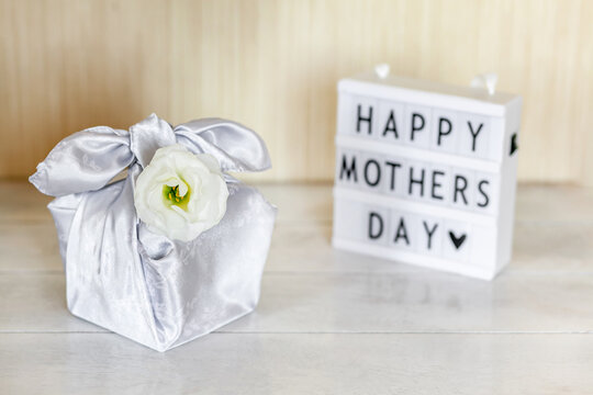 Gift box trendy wrapped in silk fabric in Furoshiki technique, light box with lettering Happy Mother's Day, white flower Eustoma or Lisianthus, wooden background. Zero Waste Life Concept