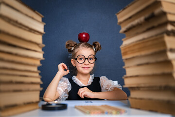 Happy smart girl in rounded glasses sitting between two piles of books with red apple on head and...