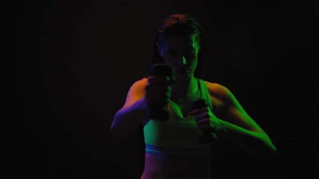 Fitness girl doing workout with dumbbells in neon lights wearing sport top stretching neck muscles turning on black background. Sport neon light. FHD footage.