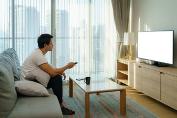 An Asian man holds a TELEVISION remote and is pressing the channel while watching TV on the couch...