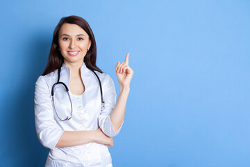 Portrait of a beautiful young doctor woman in a white robe on a blue background. A place for text and advertising. Pediatrician. Therapist. Home doctor. Doctor online