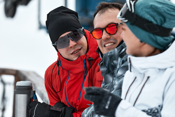 group of skiers friends on the mountain are resting and drinking coffee from a thermos on the background of the ski lift