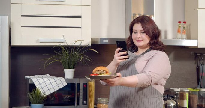 Attractive plus size woman taking pictures of delicious burger or recording video for social networks using smartphone in the kitchen. Blogging and social media concept