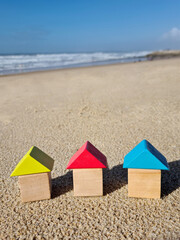 Colourful blue yellow red miniature wooden kids toy home model on sunny sand beach with blue sky, ocean background. Copy space of family lifestyle and business real estate property investment concept