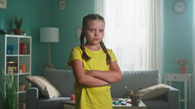 Portrait of small upset girl with two braids and green eyes, in yellow T-shirt, standing at home and looking at camera, feeling sad and depressed, Slow motion.