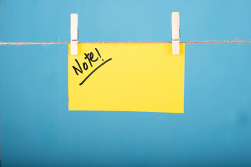 Note word written on a Yellow color sticky note hanging with a wire in front of Blue background