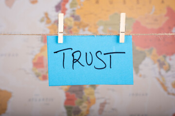 Trust word written on a Blue color sticky note hanging with a wire in front of world map background.