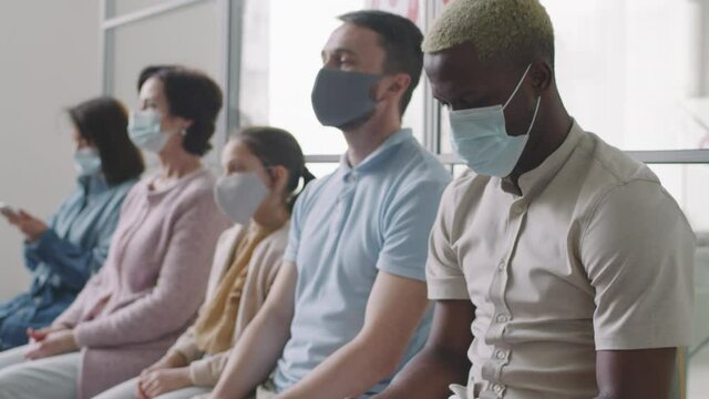 Tilting-up medium shot with slow motion of multiethnic people in protective masks sitting next to each other waiting in hospital queue before getting vaccinated