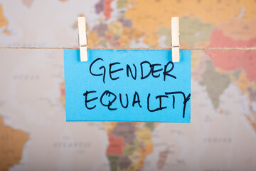  Gender Equality word written on a Blue color sticky note hanging with a wire in front of world map background.