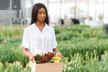 Beautiful young smiling african american girl, worker with flowers in greenhouse. Concept work in the greenhouse, flowers.