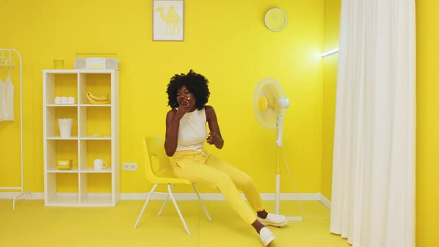 Young African Woman Is Sitting On Yellow Chair In Front Of Fan, Chewing A Bubble Gum And Laughing, Sharing Positive Emotions And Having Fun, Slow Motion.