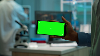 Naklejka na ściany i meble Close up of scientist woman holding horizontal phone with green mockup in modern equipped lab. Team of microbiologists doing vaccine research writing on device with chroma key, isolated display.