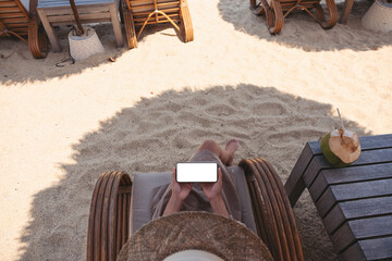 Top view mockup image of a woman holding mobile phone with blank desktop screen while sitting on the beach