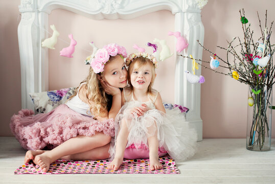 Two beautiful girls in flower wreaths and magnificent skirts sitting near fireplace. Easter, spring concept.