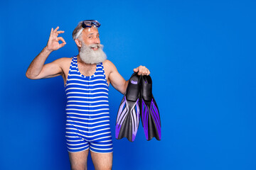 Photo of funky funny mature smiling man hold slippers showing okay sign prepare to swim isolated on blue color background © deagreez