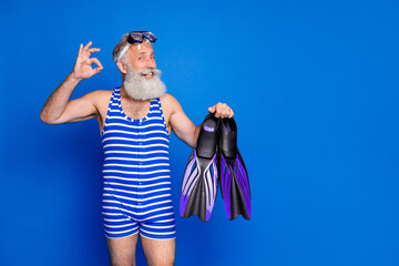 Photo of funky funny mature smiling man hold slippers showing okay sign prepare to swim isolated on blue color background
