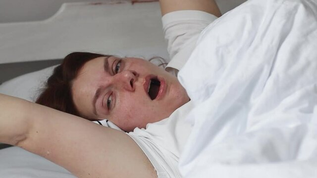 Sleepy young adult woman waking up lying in comfortable white bed. Wake up in the morning.
