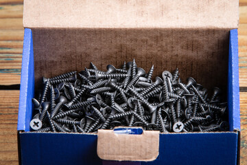 Black screws in a cardboard box. Wooden texture background. Screws for plasterboard