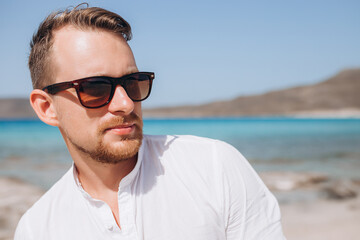 portrait of a young man in sunglasses and a white shirt against the background of the sea and rocks. Sumer holidays 