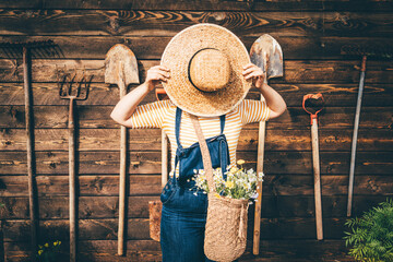 Woman wearing straw hat and blue denim dungarees relaxing near wooden old summerhouse wall on sunny...