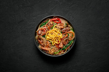 Obraz na płótnie Canvas Japchae in black bowl on dark slate table top. Korean cuisine glass chapchae noodles dish with vegetables and meat. Asian traditional food. Authentic meal. Top view