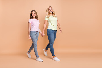 Fototapeta premium Full size profile side photo of happy woman and little girl go empty space hold hands isolated on beige color background