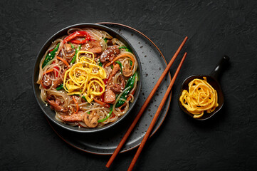 Japchae in black bowl on dark slate table top. Korean cuisine glass chapchae noodles dish with vegetables and meat. Asian traditional food. Authentic meal. Top view