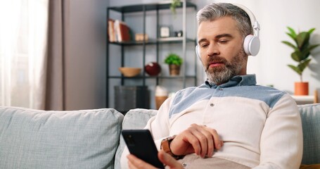 Naklejka na ściany i meble Close up of Caucasian handsome middle-aged bearded male sitting in living room at home relaxing putting earphones and listening to music from smartphone, digital gadget, leisure concept