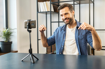 Young mature male blogger sitting at the desk, stylish apartment, recording vlog in front of mobile phone standing on a tripod, pointing down with fingers offering to subscribe to the page