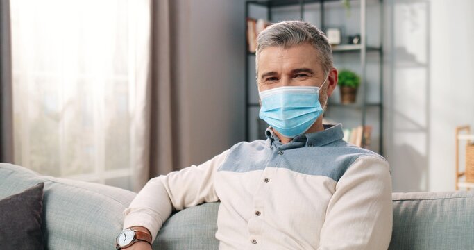 Close Up Portrait Of Caucasian Middle-aged Senior Happy Man In Medical Mask Sitting Isolated At Home In Living Room In Positive Mood, Happy Face, Resting In Apartment, Covid-19 Coronavirus Concept