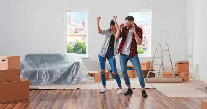 Happy cheerful young Caucasian cute married couple man and woman moving home into new house celebrating dancing and having fun getting settled together. Many carton boxes Relocation concept Moving day
