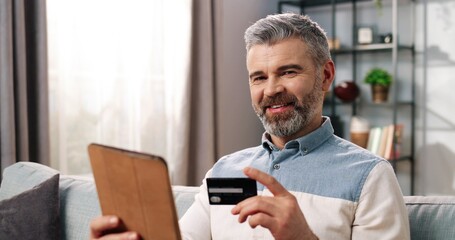 Close up portrait of cheerful Caucasian handsome middle-aged male sitting on couch in cozy room at home buying online making purchase with credit card on tablet, e-commerce, shopping concept