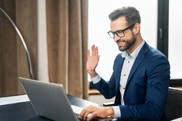 Video call concept, young good-looking man wearing suit and glasses is using a laptop for video connection with family, remote meeting, say hi, looking at the webcam, glad to see, in stylish office