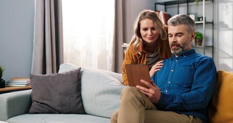 Portrait of handsome Caucasian middle-aged bearded grey-haired man sitting on sofa in room surfing internet on tablet browsing and choosing something with his beautiful happy wife, family concept - Powered by Adobe