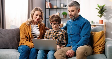 Portrait of happy Caucasian parents mom and dad with little child son sitting on sofa at home using laptop computer surfing internet choosing something online with parents, e-commerce concept