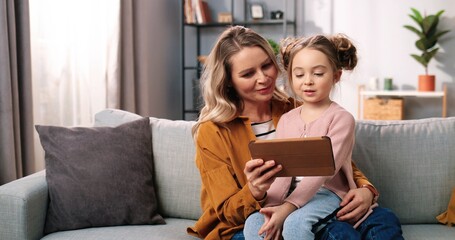 Close up portrait of happy Caucasian family mother and little cute daughter tapping on tablet device surfing internet spending time together sitting on couch in living room at home, parenting concept