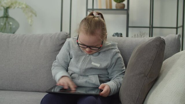Cute positive preadolescent special needs girl in eyeglasses sitting on couch, networking and browsing social media content using digital tablet while engaged in online education at home.