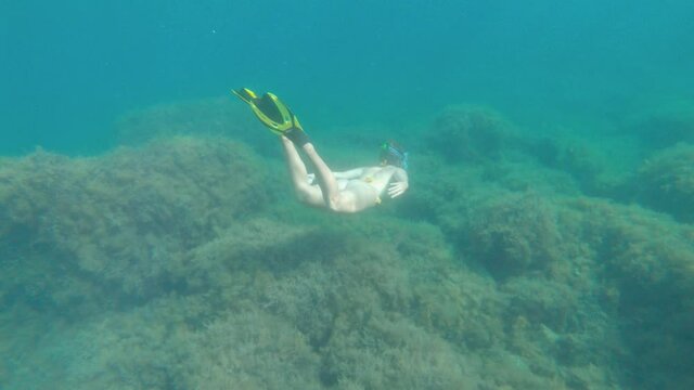 A woman in a yellow swimsuit with a snorkel mask and fins swims over a coral reef on a sunny day, shot in slow motion.